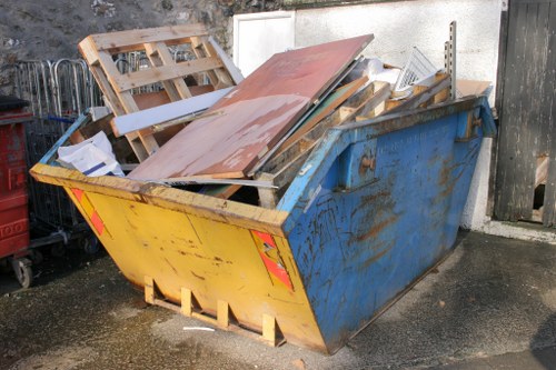 A large, rectangular industrial skip with a yellow front panel and blue sides, filled with various waste materials, is positioned on a paved surface near a white wall and a black wooden fence. The contents include wooden pallets, panels, and assorted debris, some with visible textures like rough plywood and smooth painted surfaces, predominantly in shades of brown, white, and grey. The skip appears weathered with rust and dirt marks on the exposed metal surfaces, and it is situated outdoors, likely on a driveway or service yard in Highgate, suitable for rubbish removal services by Highgate House Clearance. Surrounding objects include additional black waste bins and a segment of fencing, with natural light illuminating the scene, highlighting the contrasting textures and colors of the waste materials against the background. The environment suggests a property clearance or waste collection setting typical of residential or commercial premises in the local area.