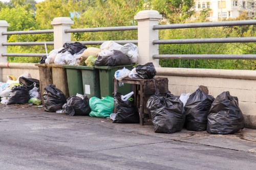 A collection of black rubbish bags, some tied at the top and others partially open, along with a few green and white bags, are piled onto the edge of a paved pathway next to a waist-high concrete barrier with metal railing posts. The bags contain various debris and household waste, with some bags leaning against the barrier and others placed directly on the ground, indicating a waste collection or clearance activity. In the background, there are trees and greenery, suggesting an outdoor setting possibly within a suburban or urban area near a park or residential street. The scene is illuminated by natural daylight, highlighting the different textures of the plastic bags and the smooth surface of the pavement. This image reflects rubbish removal or house clearance services that a company like Highgate House Clearance might provide in the Highgate area, focusing on the disposal of household waste accumulated on a street or residential driveway.