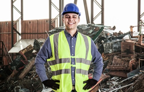 A male waste management worker wearing a blue safety helmet, a high-visibility yellow vest, a blue shirt, and black gloves stands indoors in front of a large heap of various waste materials, including broken furniture, wooden panels, and metal debris. The background shows a spacious industrial setting with metal framework and sliding doors, suggesting a waste sorting or disposal facility near Highgate. The worker is smiling confidently, and the scene is well-lit to highlight the textures of the waste and the safety gear. This image visually represents rubbish clearance services provided by Highgate House Clearance, emphasizing efficient waste removal within London suburbs such as Highgate.