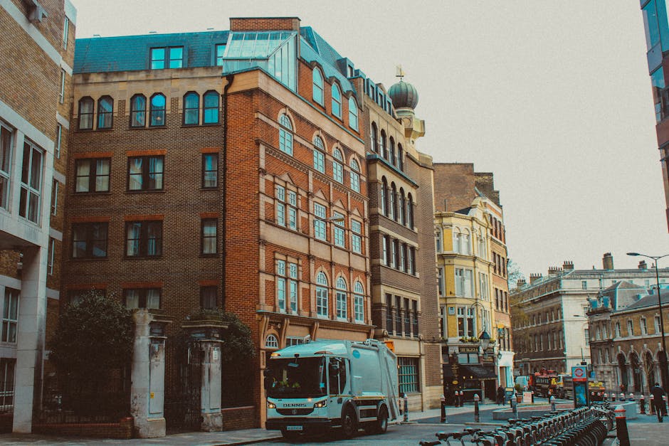 A street scene in an urban area featuring a mix of historic and modern buildings. The foreground shows a grey waste collection truck parked on the side of the street, with its cab facing left. The tru